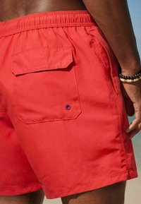 Röda simshorts i mjukt tyg, med en bakficka med lock och ögla, elastisk midja och ungefär knälång längd.