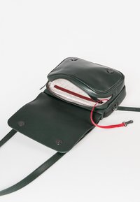 Borsa a tracolla verde in pelle con finitura liscia, scomparto principale con zip e un interno decorativo con tirazip rosso. Dettagli con chiusura a bottone.