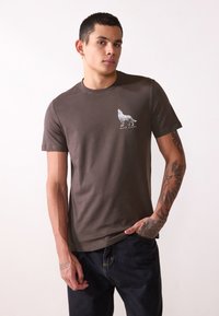 Bruine katoenen T-shirt met een ronde hals, voorzien van een witte wolfafbeelding en de tekst "WALK ALONE" eronder. Korte mouwen, ontspannen pasvorm.