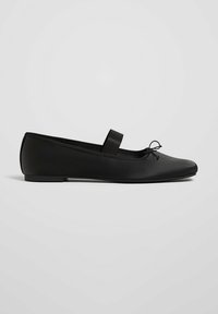 Ballerine plate en cuir noir avec une fine lanière sur le dessus et un petit nœud près de l’orteil arrondi, semelle plate.