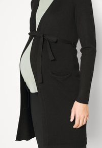 Černý pletený kardigan s páskem na zavazování, dlouhými rukávy a dvěma bočními kapsami, nošený přes světle zelený žebrovaný top.