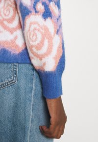 Sweter o niebieskiej bazie, z przewymiarowanymi wzorami róż w kolorze różowym i białym. Teksturowane rękawy z dzianiny z prążkowanymi mankietami, w połączeniu z dżinsami.