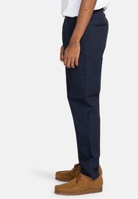 Navy blue cotton trousers with a straight leg fit, side pockets, and a back pocket. Worn with brown suede moccasin shoes.