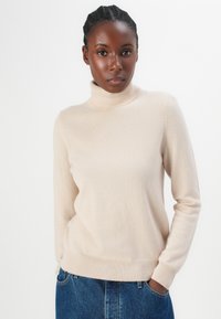 Lys beige høyhalset genser laget av myk strikket stoff. Lange ermer, ribbestrikkede mansjetter og kant. Kombinert med blå denimjeans.
