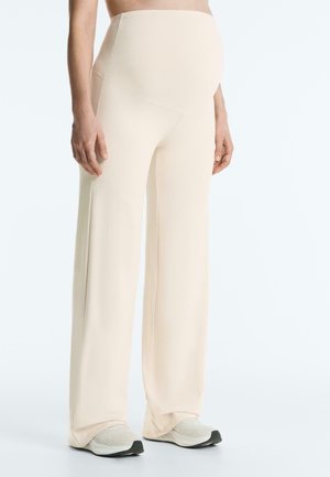 Gravid person iført beige graviditetsbukser med højt liv og vide ben samt hvide sneakers, stående foran en ensfarvet baggrund.