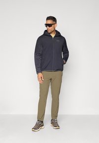 Veste noire zippée avec capuche et poches latérales, pantalon vert olive, et chaussures de randonnée grises avec accents beiges. Lunettes de soleil foncées portées.