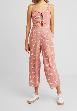 Vrouw draagt een mouwloze jumpsuit in rozenroze met bloemenprint, met verbonden voorzijde en openingen, en witte platform sneakers, staand tegen een effen achtergrond.