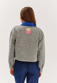 Šedý skrátený mikina s modrým rebrovaným golierom a ružovým logom na chrbte. Mäkká textúra, dlhé rukávy s gombíkovými manžetami.