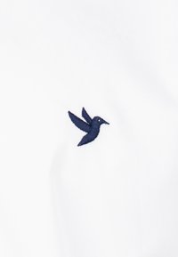 La tela blanca presenta un pequeño logo de colibrí bordado en azul marino en el lado izquierdo. La textura es suave y lisa.