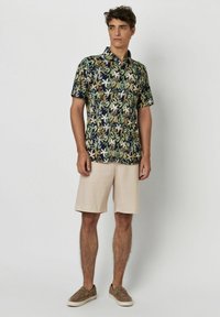 Korte mouwen marineblauw shirt met een meerkleurig tropisch bladerenpatroon, gecombineerd met beige shorts en bruine instappers.