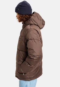 Hnedá izolovaná puffer bunda s kapucňou, ktorá má zipsovú kapsu, elastické manžety a hladký, lesklý povrch.