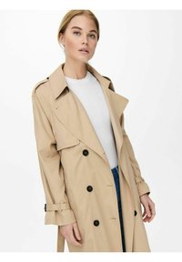 Beige trenchcoat med dobbeltradet design, store revers, svarte knapper og belte ved ermet, båret over en hvit skjorte.