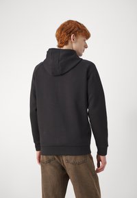 Felpa nera con cappuccio, con polsini e orlo a costine, texture in felpa e maniche raglan, indossata con pantaloni chiari in tessuto strutturato di colore marrone.