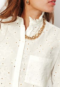 Blouse blanche à boutons avec motifs floraux brodés et découpes en œillet. Col volanté, poche poitrine, et collier chaîne dorée en accent.