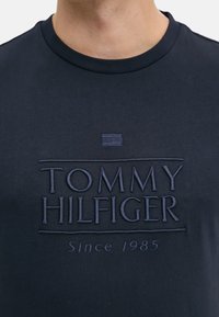 T-shirt in cotone blu navy con logo ricamato in rilievo "TOMMY HILFIGER" e la scritta "Dal 1985" sottostante. Collo rotondo e maniche corte.