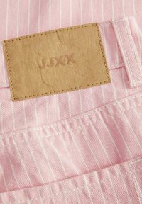 Roze prugasta tkanina s teksturiranim kožnim gumbom, utisnutim s "JJXX." Vidljivi detalji šivanja oko gonga i tkanine.