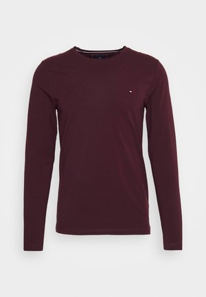 Langærmet, mørk burgundy crewneck-skjorte med lille rød, hvid og blå logo på venstre bryst, vist på en neutral baggrund.