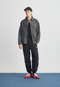 Fekete mintás dzseki, fekete póló, fehér csíkos fekete tréningnadrág és piros sportcipők. Egy lapos karimájú fekete sapka egészíti ki a megjelenést.