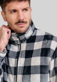 Chaqueta de forro polar con un patrón de cuadros en blanco y negro, que presenta un cuello alto, cremallera frontal y una textura suave.