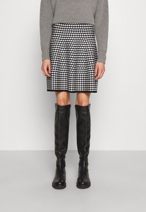 Osoba ubrana w szary sweter, czarno-białą spódnicę w kratę jodełkę oraz czarne kozaki do kolan, stojąca na tle jednolitego białego tła.