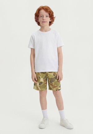 Παιδί με κόκκινα μαλλιά όρθιο, φορά απλό λευκό t-shirt, σορτς με καμουφλάζ σχέδιο, λευκές κάλτσες και λευκά αθλητικά παπούτσια, μπροστά σε λευκό φόντο.