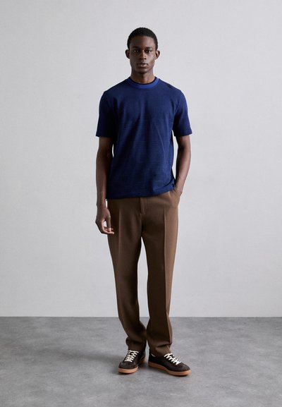 Marineblå t-skjorte med korte ermer i et teksturert stoff, kombinert med brune skreddersydde bukser og mørke sneakers med lys såle og hvite lisser.