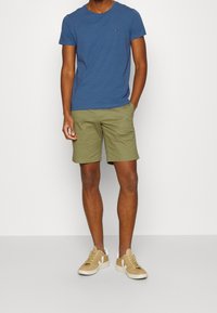 Blue cotton t-shirt, olive green shorts, and beige sneakers. T-shirt has a small logo; shorts are straight-cut with pockets.