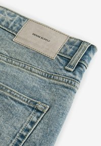Jeans en denim bleu clair avec une surface texturée, présentant une étiquette beige embossée avec "DENIM SUPPLY" et des détails de couture standard.