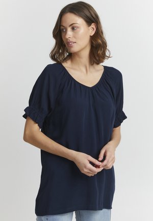 Navyblaues kurzes Oberteil mit V-Ausschnitt, gerafftem Halsauschnitt und Rüschensäumen. Weicher Stoff mit lockerer Passform, kombiniert mit Jeans.
