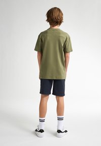 Camiseta de manga corta color verde oliva, combinada con pantalones cortos azul marino, calcetines a rayas en blanco y negro, y zapatillas negras con suelas blancas.