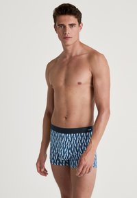 Boxershorts hebben een marineblauwe basis met een herhalend wit en groen golfpatroon, gemaakt van een zachte, rekbare stof. Inclusief elastische tailleband.