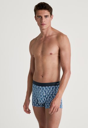 Die Boxershorts bestehen aus einem marineblauen Grundton mit einem sich wiederholenden Muster aus weißen und grünen Wellen, gefertigt aus einem weichen, dehnbaren Stoff. Mit elastischem Bund.