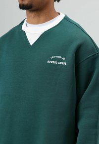 Groene sweatshirt met een v-hals design, met een witte kraaglaag eronder. Aan de voorkant staat witte tekst: "LE-75005-DE STUDIO ABYSS."