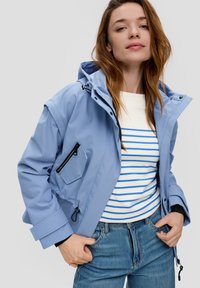 Jasnoniebieska wodoodporna kurtka z kapturem, zapinana na zamek, z boczną kieszenią i elastycznymi mankietami, noszona na biało-niebieską w paski koszulę oraz dżinsy.
