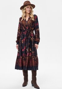 Długa sukienka z długim rękawem w kwiatowy wzór na ciemnej tkaninie, z talią podkreśloną paskiem, falbaną na dole i mieszanką kolorów czerwonego, złotego oraz ciemnozielonego. Brązowe botki i kapelusz.