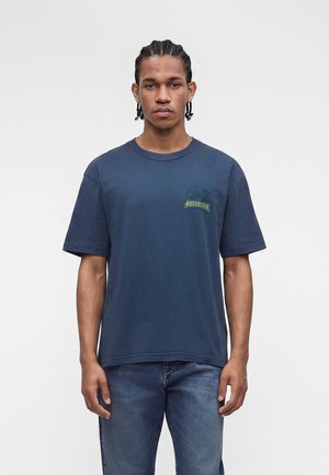 Νέος άνδρας με πλεξούδες που φοράει μπλε μπλούζα κοντομάνικη με ένα μικρό γραφικό στο στήθος και μπλε τζιν.