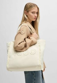 Borsa tote in tela bianca con scritta "HUGO" in rilievo, bordi sfrangiati e rifinitura morbida, portata da una persona con una maglietta beige e denim.