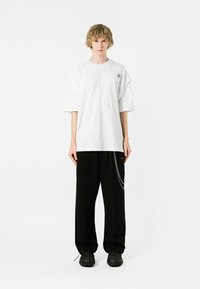 Camiseta blanca de gran tamaño con mangas cortas, que presenta un pequeño logo y detalles de corte. Combinada con pantalones holgados negros y calzado negro.
