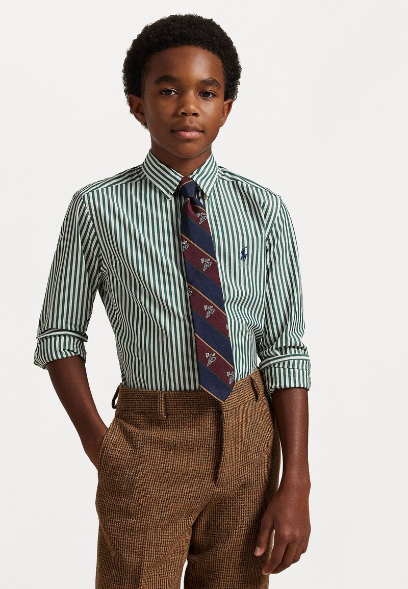 Zelena in bela črtasta srajca s zavihanimi rokavi, v kombinaciji z rjavimi vzorčastimi hlačami in mornarsko kravato z rdečimi poudarki.