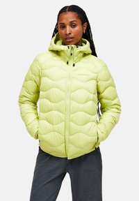 Lys grøn pufferjakke med quiltet bølgedesign, høj krave, lynlåslukning foran, hætte med snoretræk, sorte detaljer, båret over mørke teksturerede bukser.