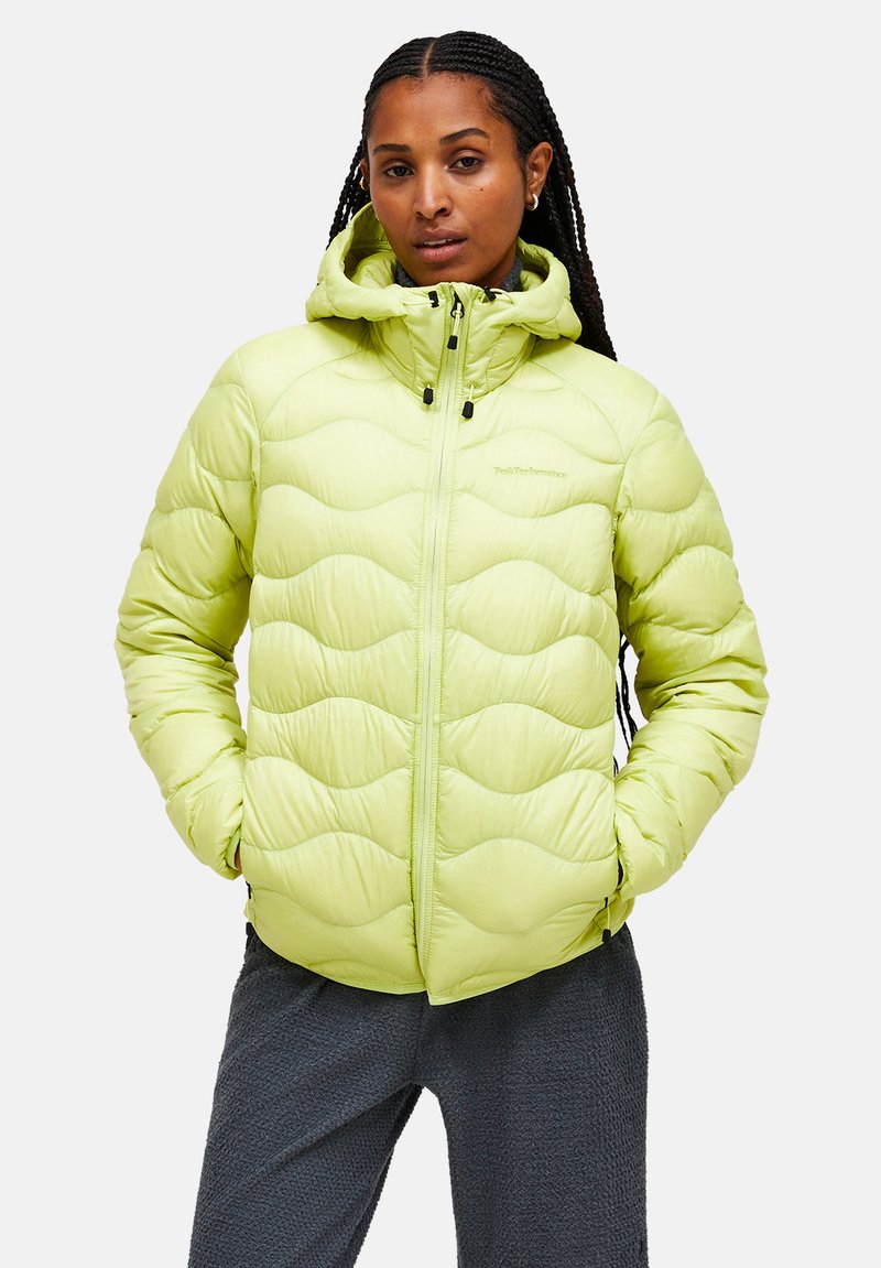 Lys grøn pufferjakke med quiltet bølgedesign, høj krave, lynlåslukning foran, hætte med snoretræk, sorte detaljer, båret over mørke teksturerede bukser.