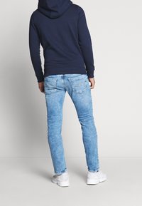 Mężczyzna ubrany w granatowy hoodie, jasnoniebieskie dżinsy i białe sneakersy, stojący plecami na tle jednorodnym w jasnym kolorze.