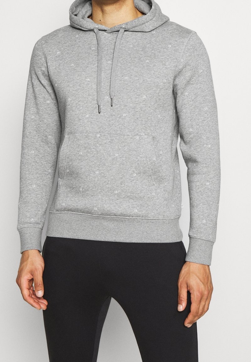 Homme portant un sweat à capuche gris clair avec de petits logos blancs "ck" et un pantalon noir, debout contre un fond uni.