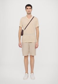 Beige T-Shirt mit kurzen Ärmeln und V-Ausschnitt aus strukturiertem Stoff, kombiniert mit hellbeigen Shorts. Das Model trägt eine kleine braune Umhängetasche. Weiße Sneakers.