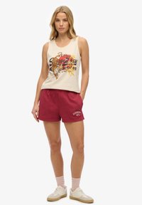 Vrouw draagt een mouwloos beige top met tijgerprint en rode korte broek met zijzakken, staand tegen een witte achtergrond.