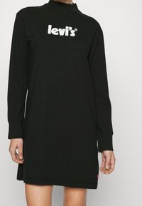 Personne portant une robe noire à manches longues Levi's avec un logo blanc sur la poitrine et une étiquette rouge près de l'ourlet.