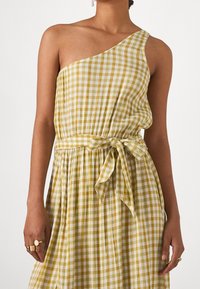 Eén-schouder jurk in een geel en wit ruitpatroon. Heeft een aansluitende taille met een gestrikte riem, vloeiende stof en een ontspannen silhouet.