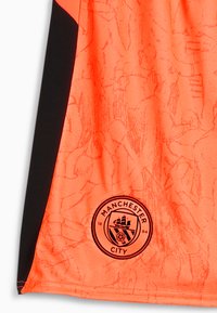 Camiseta deportiva de color naranja brillante con un patrón texturizado, que presenta acentos laterales negros y un logo negro de Manchester City en la parte inferior.