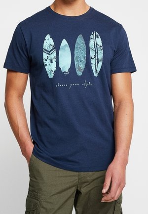 Mężczyzna noszący granatowy T-shirt z czterema stylizowanymi, jasnoniebieskimi deskami surfingowymi i napisem „wybierz swój styl” na piersi.