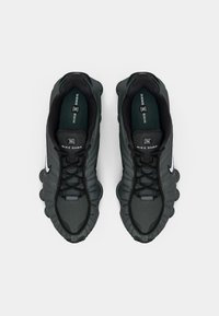 Juodos Nike Shox sportbačiai, turintys tinklelio viršutinę dalį, tekstūruotą apdailą, paminkštintą apykaklę ir išskirtinį paminkštinto padažo dizainą.